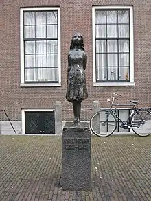 A bronze statue of a smiling Anne Frank, wearing a short dress and standing with her arms behind her back, sits upon a stone plinth with a plaque reading "Anne Frank 1929–1945". The statue is in a small square, and behind it is a brick building with two large windows, and a bicycle. The statue stands between the two windows.