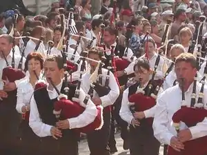 Group of young men and women, wearing white shirts (some with black waistcoats) and black trousers, marching in a parade, in the sunshine. Each is playing a bagpipe. The bag is a claret colour. The entire picture is full of people. Those not taking part in the parade are watching the procession.