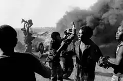 A group of men standing in front of an overturned car and a plume of black smoke.