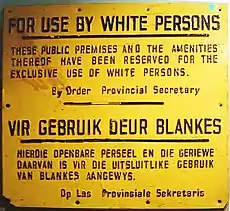 A rectangular yellow wall plaque. It contains a message written in black in both English and Afrikaans. In English, the message reads: "For Use by White Persons. These public premises and the amenities thereof have been reserved for the exclusive use of white persons. By Order Provincial Secretary".