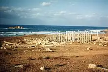 Several columns standing in a brown landscape, in front of the sea. An island is visible.