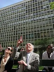 Photograph showing a man pointing upwards to something behind the photographer. The man is elderly, and is flanked by three younger people. There is a large, multistory building behind the people.