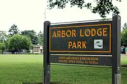 Wooden sign with text identifying the name, management, and hours of operation for the park; in the background is a green lawn, part of a building, and two people.