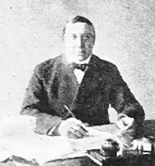 Black and white portrait of a man seated behind a table, with a pen in his right hand.