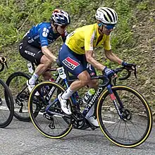 Two women on bicycles. One wearing a yellow top and blue shorts, the other wearing a blue top and black shorts