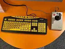 An assistive keyboard with bright yellow, large print keys. On its right, there is a joystick that is used as an alternative mouse. The keyboard and joystick are on a table that is located in Punggol Regional Library.