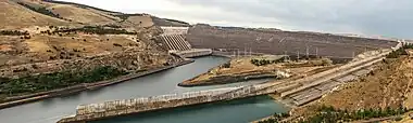 Two large channels of water flow slowly from a low sloping dam with several pipes at one end of the dam