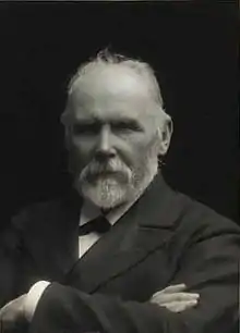 Black and white portrait of a bearded man with folded arms, wearing a suit