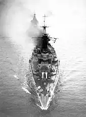 Photograph looking down on a large warship. The ship is travelling at speed, and is about to lead two or three other large vessels under the photographer.