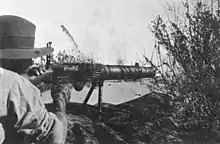 A soldier lies among the grass in the prone position behind a machine gun which he is holding in the shoulder.