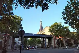 The pedestrian bridge of the "Promenade Plantée" ("Coulée Verte") traverses the avenue at the intersection of Avenue Daumesnil. The steeple of St. Antoine church is also visible.