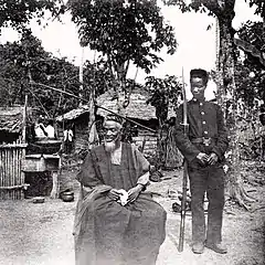 Image 24Temne leader Bai Bureh seen here in 1898 after his surrender, sitting relaxed in his traditional dress with a handkerchief in his hands, while a Sierra Leonean West African Frontier Force soldier stands guard next to him  (from Sierra Leone)