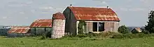 A short silo in the centre of the photograph is slanted slightly to the right, topped by a conical red roof. Three barns form a V shape behind the silo. To its right is a large barn, with slanted red roof and open doors. Perpendicular to it are two similar but smaller barns in series, visible to the left of the silo.