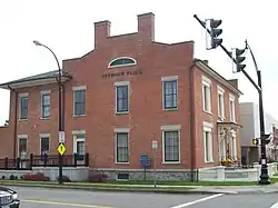 A brick building on a corner with a traffic signal in front of it. Two chimneys rise from a section that rises above the roofline on the side facing the camera; the front faces the right side. Metal lettering on the section facing the camera spells out "Seymour Place".