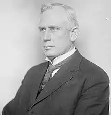 A white-haired man, sitting, wearing dark jacket, patterned tie, and white shirt