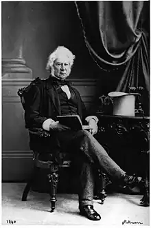 Formal portrait of a white-haired, light-skinned man sitting in a chair, wearing mid-Victoria clothing