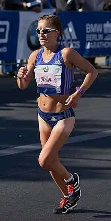 A woman wearing blue athletics running kit and sunglasses is running along a street. She has a competitor identifier 'Dixon' pinned on her chest, with the event sponsor's logo.