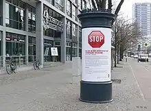 A sidewalk, bordered on the left by a modern building featuring the inscription "Scientology-Kirche" above its entrance and on the right by a cycle path, beyond which there is a road with a yellow "H" sign marking a bus stop. The pavement is lined by trees on the side facing the cycle path. In the foreground there is an advertising column with a white poster featuring a large red Stop sign, followed by text commenting on the activities of Scientology in the local area. A corner of the roof of a bus shelter is visible protruding behind the column.