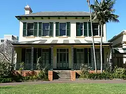 A two-storey house in the Georgian style. It has a verandah supported by openwork flat iron piers, a central door between four French windows, and five sash windows above. All the windows have shutters. The colour-scheme is white with dark green. Although simple, it is finely detailed. The garden has hardy shrubs and two tall palms to one side.