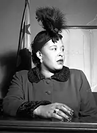 A dark-skinned woman is sitting behind a wooden desk or a counter, facing the camera and looking to the right. She is wearing a winter coat, a hat and large shining earrings. Her right hand is on the desk and there is a thick, shining ring in its ring finger. Behind her on the right hangs a flag with one darkly colored star visible.