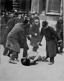 A woman is on the ground with her gloved hands over her face. A man in a top hat is holding back one police officer, while another is holding one of his gloves and stooping over the woman. In the background are several police officers and other men. Beyond them are the walls and doorway to the Parliament buildings.