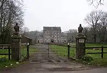 fenced and gated driveway leading through grassy area to large two and four storey building in the distance