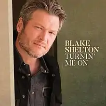 A color photograph of Blake Shelton grinning at the camera; he stands in front of a green wall.