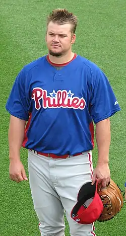 A man with spiked hair and a goatee wearing a blue baseball jersey with "Phillies" in red across the front and white baseball pants with red pinstripes stands on a baseball field holding a tan baseball glove and a red baseball cap in his left hand.