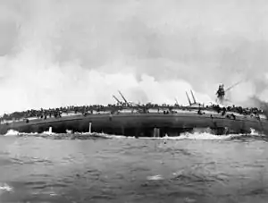 The burning hull of a ship on its side; water pours from holes punched by enemy projectiles. The keel is splitting from the intense strain.