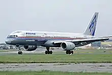 Side view of silver twin-engine jet taxiing on runway, with deployed flaps and "757" markings on tail.