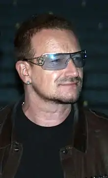 A man with facial hair wearing a leather jacket, a black shirt, an earring, and tinted glasses with a star along the frame.