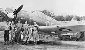 Six men in front of a single-engined military monoplane parked on a jungle airfield