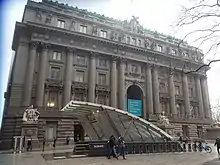 An entrance to the station in Bowling Green Park, with a glass canopy above it. Behind the entrance is the Alexander Hamilton U.S. Custom House.