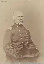 Sepia toned photo shows a seated man with a bushy moustache. He wears a dark military uniform and holds his hat in his lap.