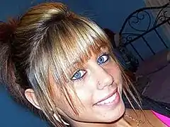 A slightly tilted view of a young white woman with blonde hair and blue eyes, with a wrought-iron bed frame in the background