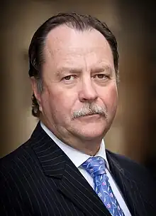 A Caucasian male in his late fifties wearing black pinstripe suit, white shirt and blue decorated tie.