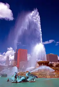 A fountain spouting water from several points with several tall buildings and a blue sky in the background.