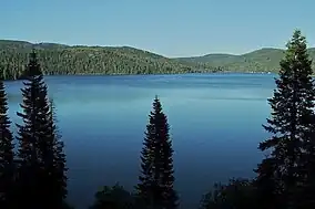 A large lake under blue skies surrounded by forested hills with steep slopes under a clear blue sky