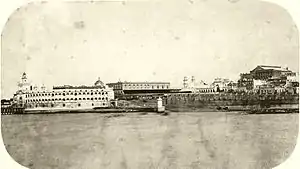 Photograph depicting a waterfront with stone quays, a paved ramp leading down to the water's edge, a fortress on the left edge of the picture, and spires, domes and multi-story buildings in the background