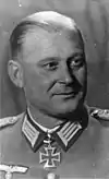 The head of a man, shown in semi-profile. He wears a military uniform with a military decoration in shape of an iron cross displayed at the front of his shirt collar. His hair is combed to back.