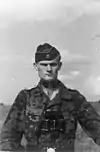 A man wearing a camouflage military uniform, side cap and a pair of binoculars around his neck. His cap has an emblem in shape of a human skull and crossed bones.