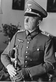 The head and shoulders of a man sitting, shown in semi profile. He wears a beaked cap and a military uniform, and an Iron Cross displayed at the front of his uniform collar. His left hand is gloved and he is holding the hilt of a saber in both hands. His facial expression is a determined; his head is pointed to the right of the camera.