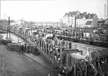 Several trains loaded with machinery take up the center of the photo. A group of nine men stand to the left.