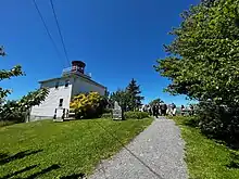 A photo taken in 2022 of the replica lighthouse (built in 1994) at Burntcoat Head Park, East Hants, Nova Scotia. The square, two storey wooden lighthouse is painted white, with a hexagonal room at the top painted red, which would have housed the light to guard ship traffic in the Cobequid Bay during the age of sail. There is a group of people walking on a pathway next to the lighthouse, and trees lining the path within the public park.