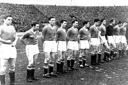 A black-and-white photograph of a football team lining up before a match. Eleven men in old-fashioned association football attire stand in a line: ten wear dark shirts, white shorts and black socks, and the other wears a still darker shirt. Behind the players can be seen one of the enormous open stands of an East European-style soccer stadium, filled to the brim with spectators.