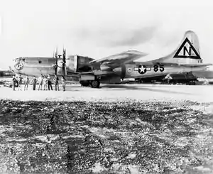 A shiny metal four-engined aircraft stands on a runway. The crew pose in front of it.