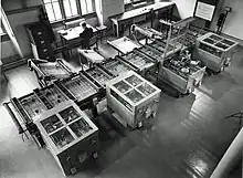 Looking down from above at a room at an intricate mechanical device which fills the room. In the background, a man sits at a desk next to a filing cabinet.Owens 1991, p. 15.