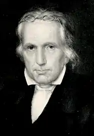 A middle-aged, studious looking man with white hair and old-fashioned glasses pushed up over his head. He is looking almost directly towards the viewer, with his shoulders turned slightly towards the left. He is wearing a dark coat and a white shirt with a broadly spread collar.