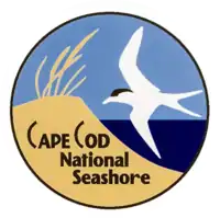 A circular logo depicting a sand dune topped with beach grass next to the ocean, with a white sea bird flying overhead.