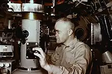 Captain Edward L. Beach dressed in khaki uniform gazing through the viewfinder while adjusting the focus using dial control on the periscope in the conning tower of the nuclear submarine USS Triton